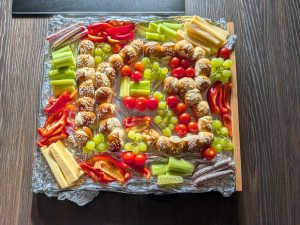 Häppchenplatte mit Obst, Gemüse und Würstchen. In der Mitte ist ein großes K3 aus kleinen Partybrötchen gelegt.